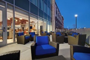 Un espace extérieur aménagé avec des meubles en osier et des coussins bleus, situé à côté d’un bâtiment doté de grandes fenêtres et d’une façade en briques.