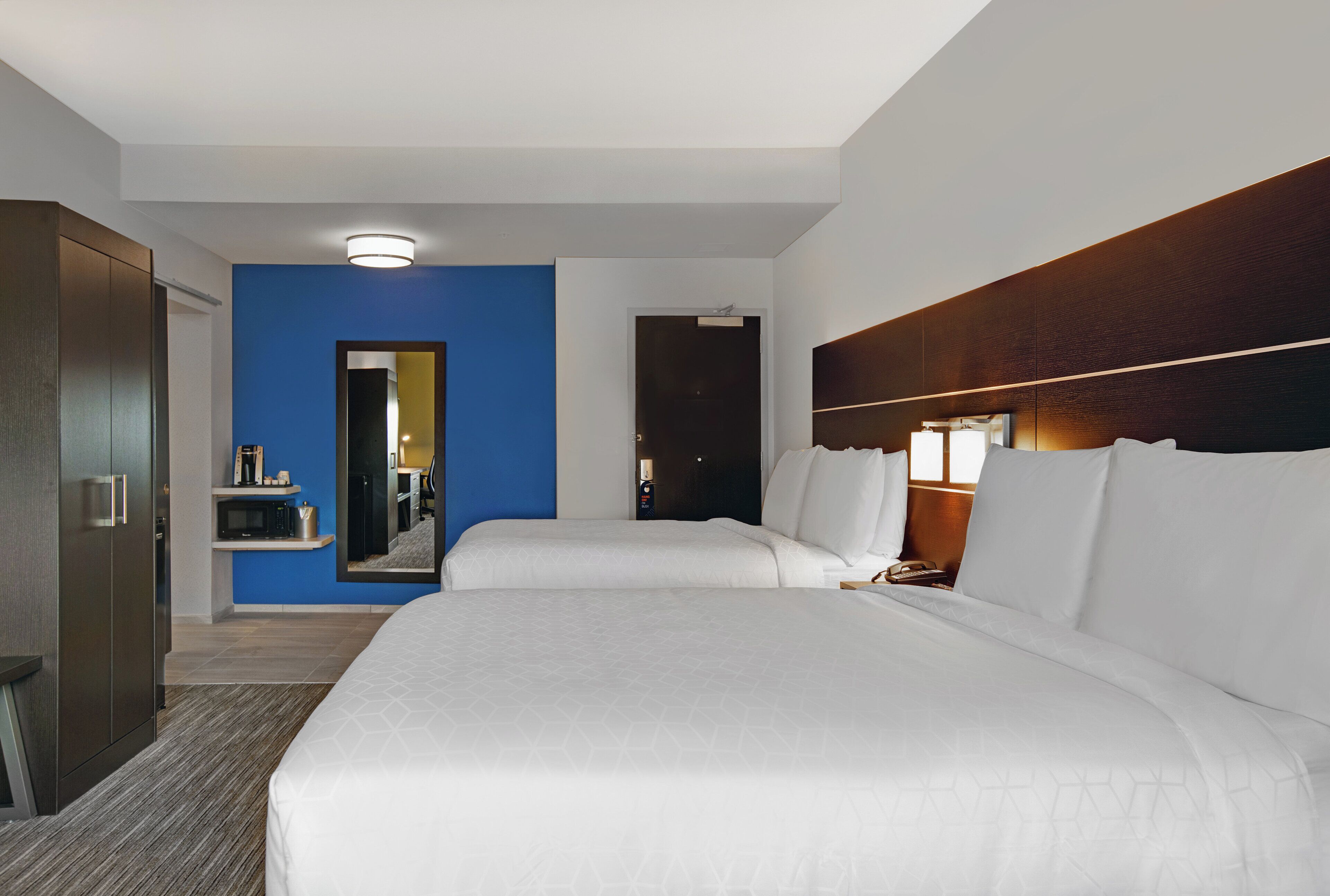 Habitación de hotel con dos camas, un armario, un espejo, una mesita de noche con una lámpara y un pequeño estante con objetos.