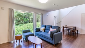 A modern living room with a blue sofa, a small round table, and a dining area with a wooden table and chairs. Large windows offer a view of greenery outside. - Wapura Rusutsu (Rusutsu)