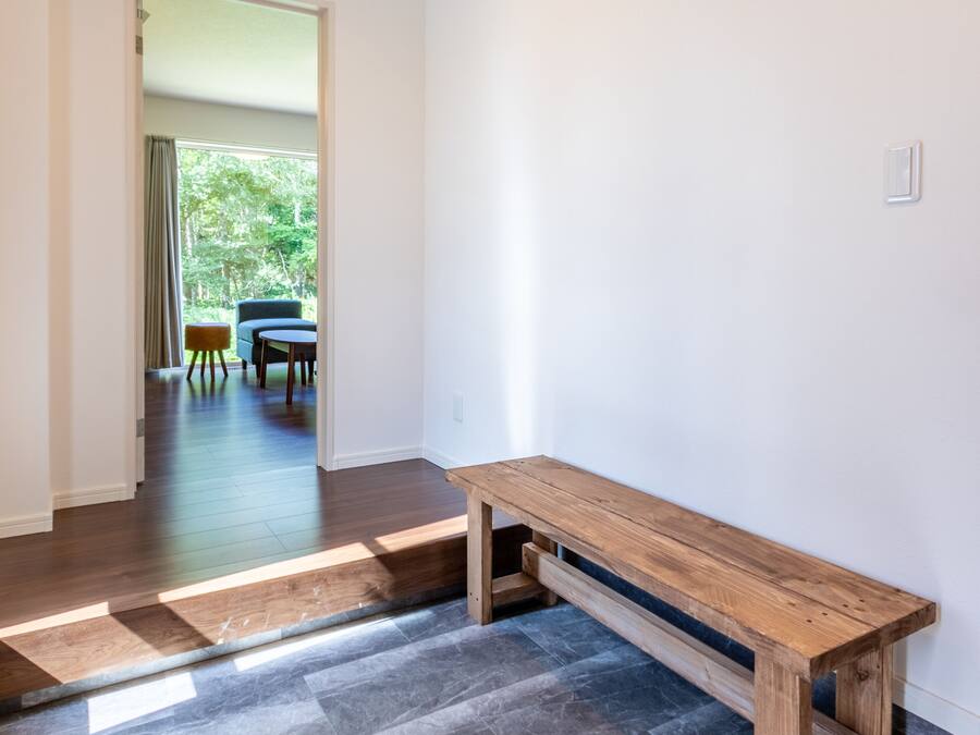 Un pasillo con un banco de madera, una puerta que da a una habitación con mesa y sillas, y vistas de follaje exterior.