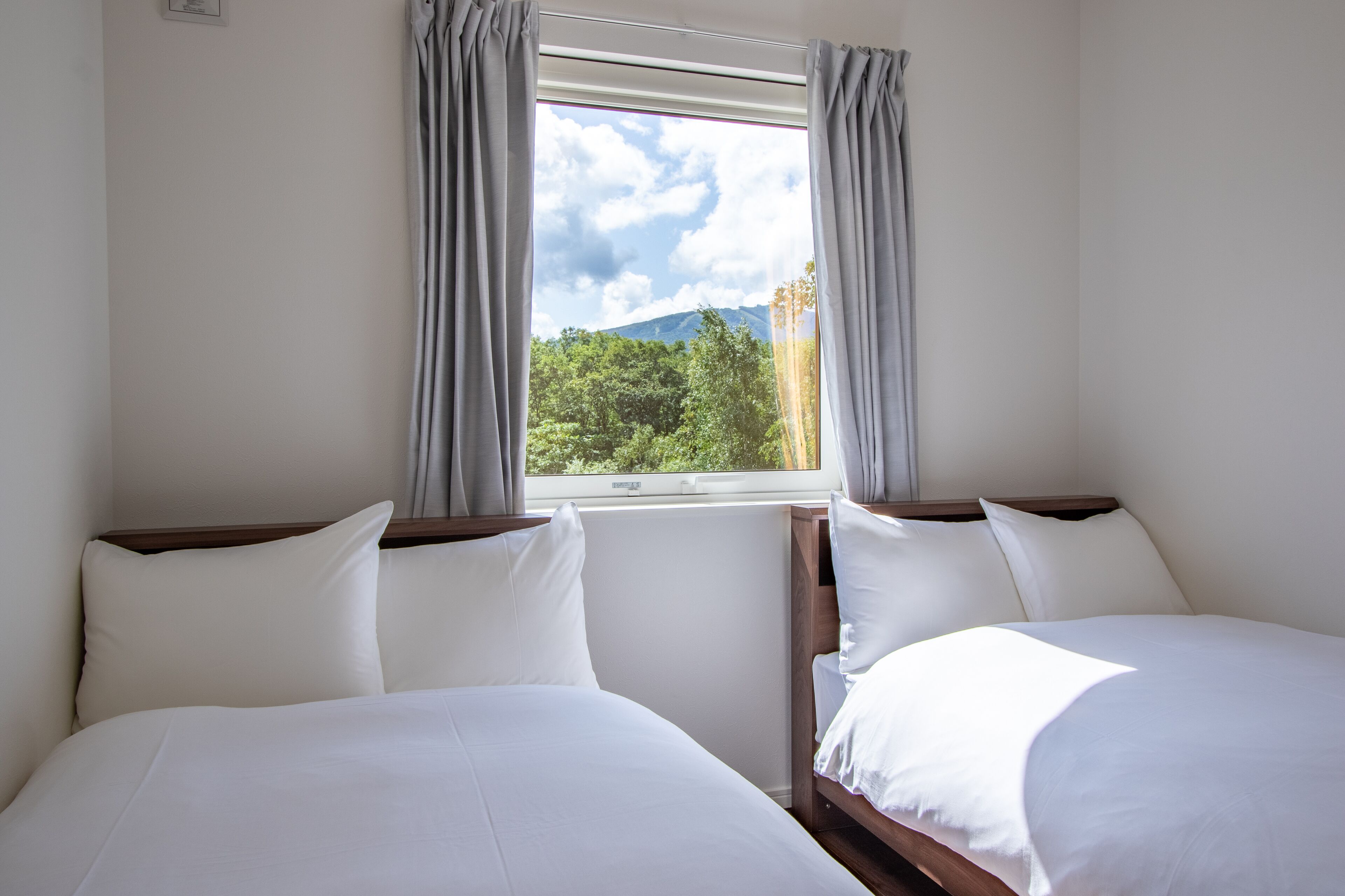 A hotel room with two beds, a window with curtains, and a view of greenery and mountains.