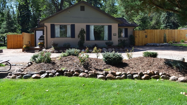 A single-story house with a sloped roof, a front porch, and a well-maintained garden with various plants and a rock border.
