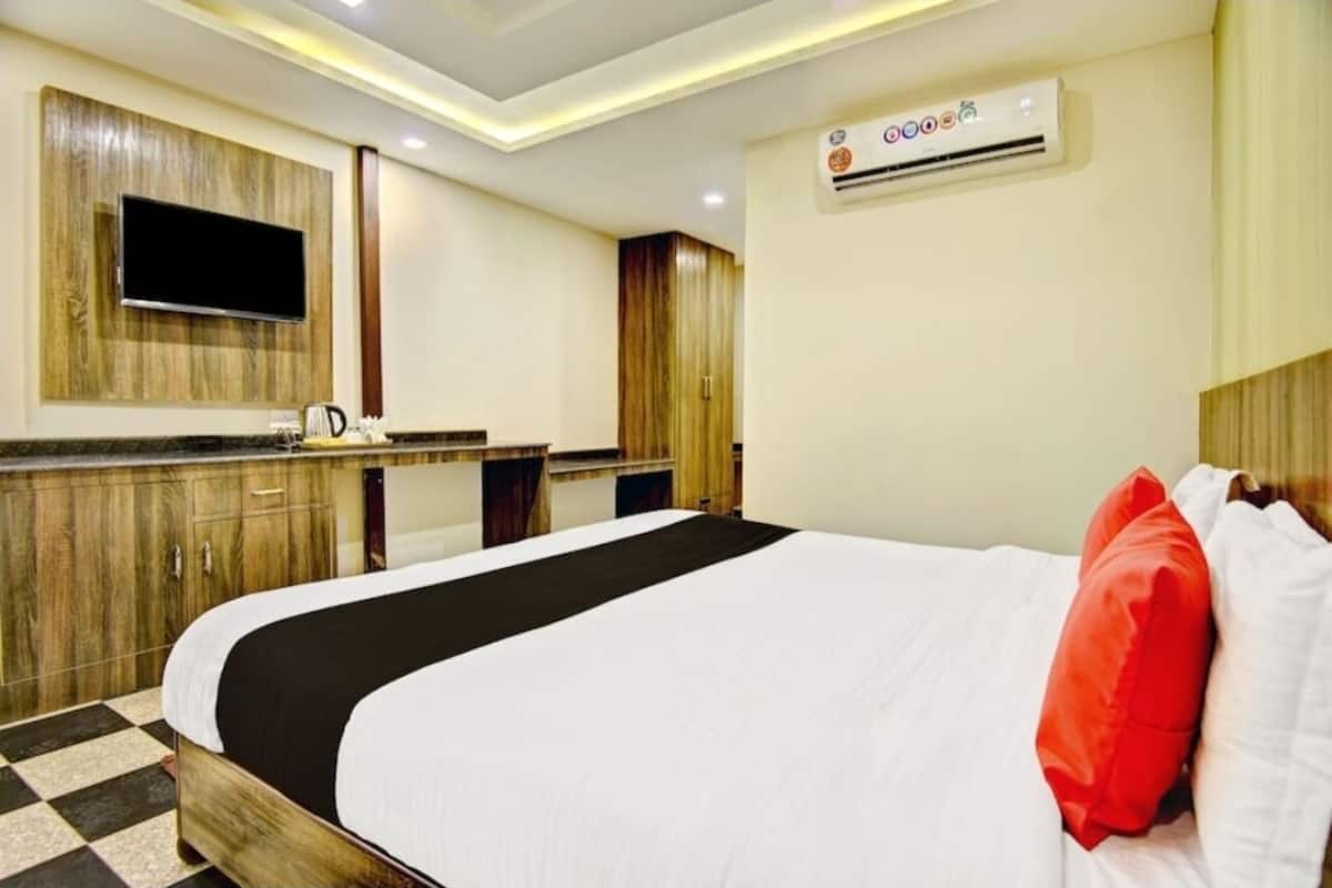 A hotel room with a bed, a flat-screen TV mounted on the wall, a wooden wardrobe, and an air conditioning unit.