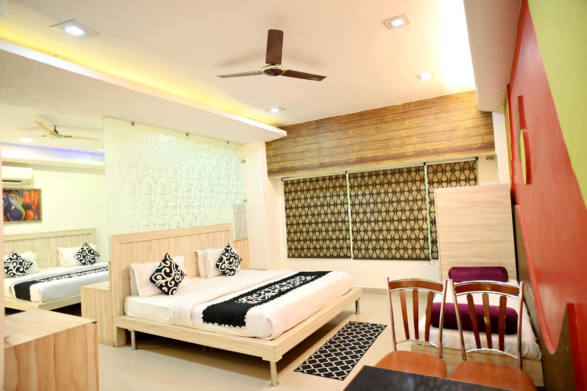 A hotel room with a bed, two bedside tables, a chair, a ceiling fan, and a window with patterned curtains.