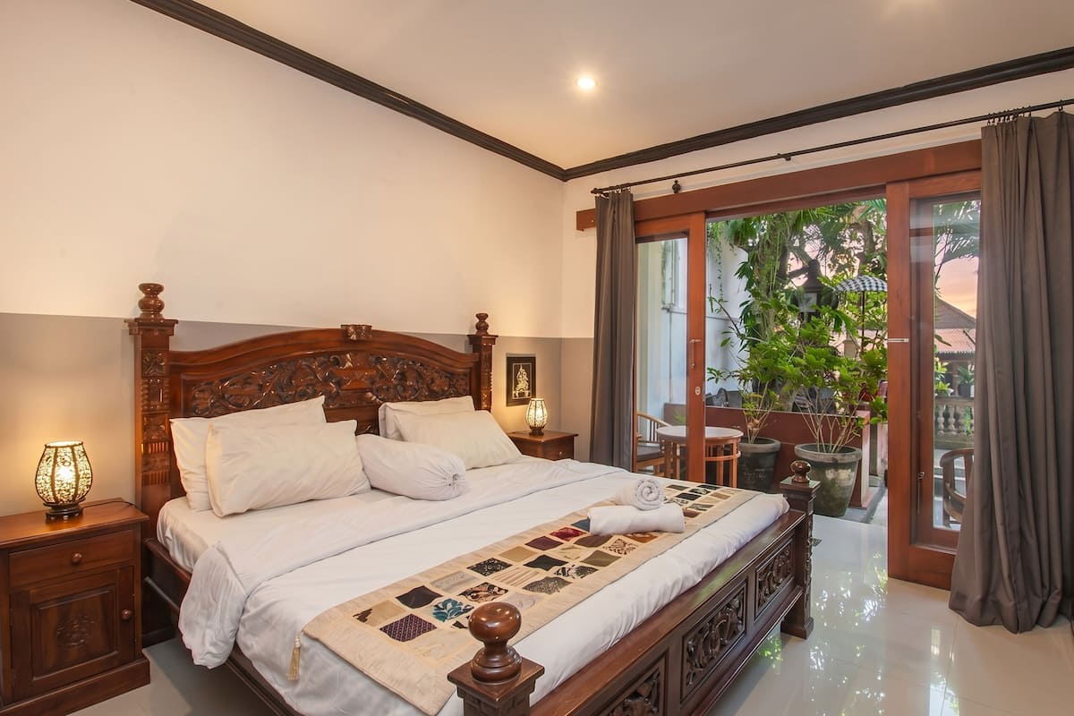 A bedroom with a large bed, a nightstand, a lamp, a view of the outdoors, and a balcony.