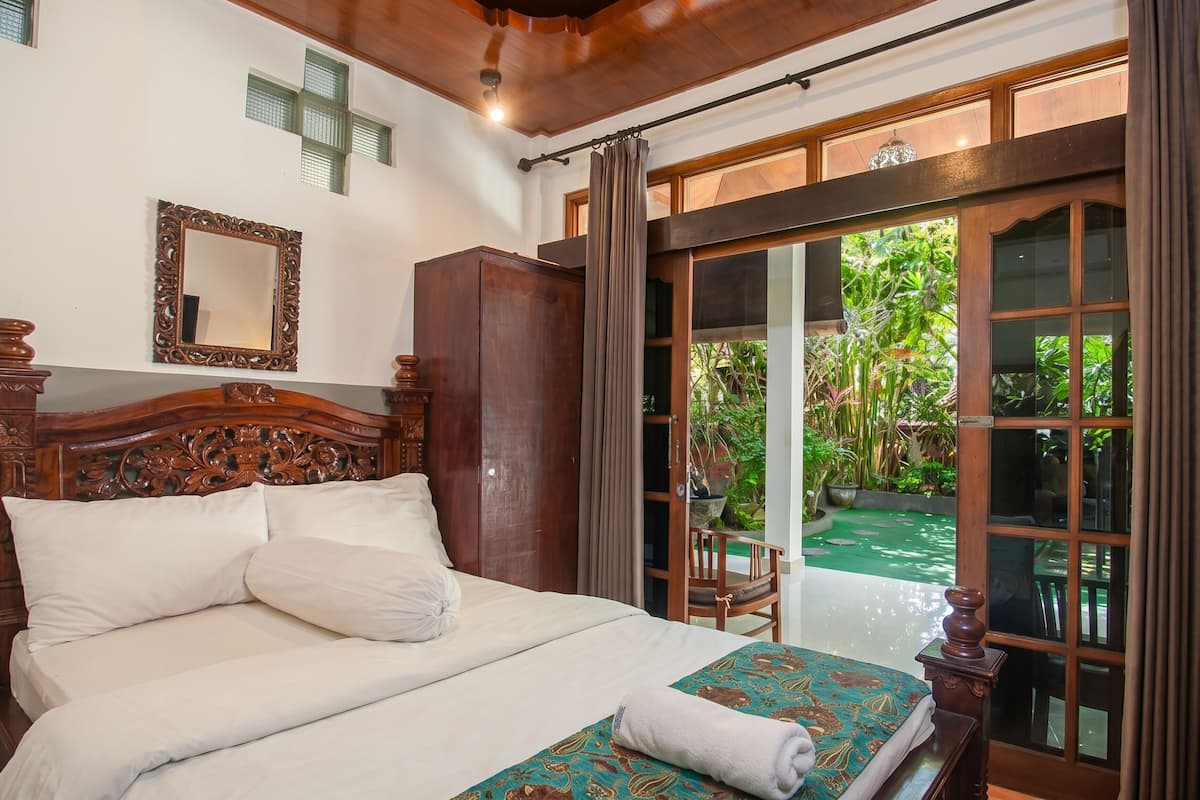 A wooden bed with white bedding, a mirror, and a view of a garden through a sliding glass door.