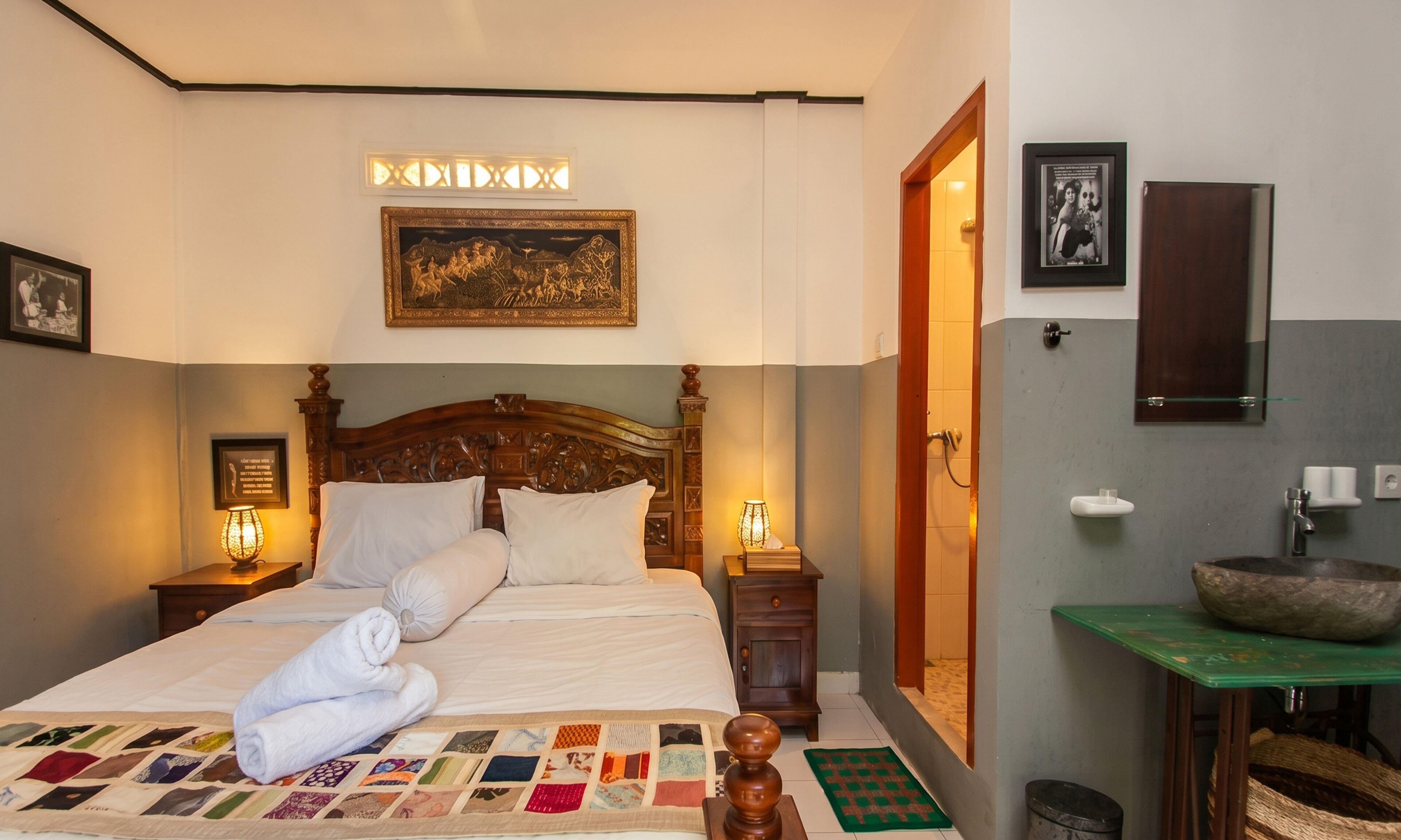 Un lit bien fait, recouvert d’une courtepointe, avec une tête de lit en bois, des lampes de chevet, un tableau encadré au-dessus du lit, une salle de bain avec un lavabo et un miroir, et une table verte avec un bol.
