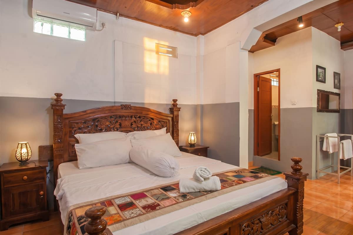 A bedroom with a wooden bed, a quilt, a nightstand with a lamp, a bathroom with a towel rack, and a wooden wardrobe.