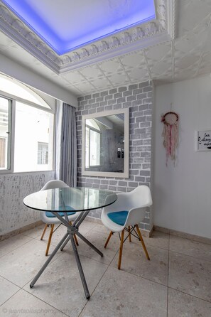 A modern room with a glass table, two white chairs, a brick-patterned wall, a mirror, and a dreamcatcher.