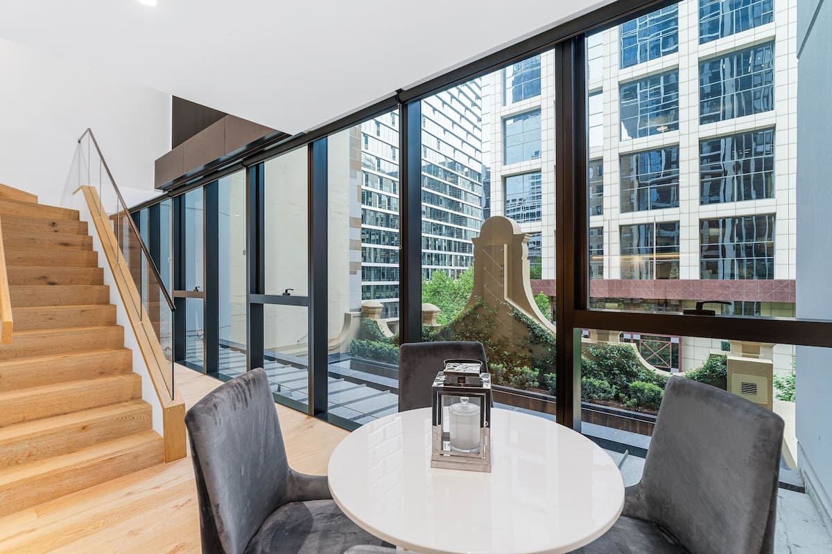 A modern interior with a staircase, a round table with chairs, and large windows offering a view of a cityscape.