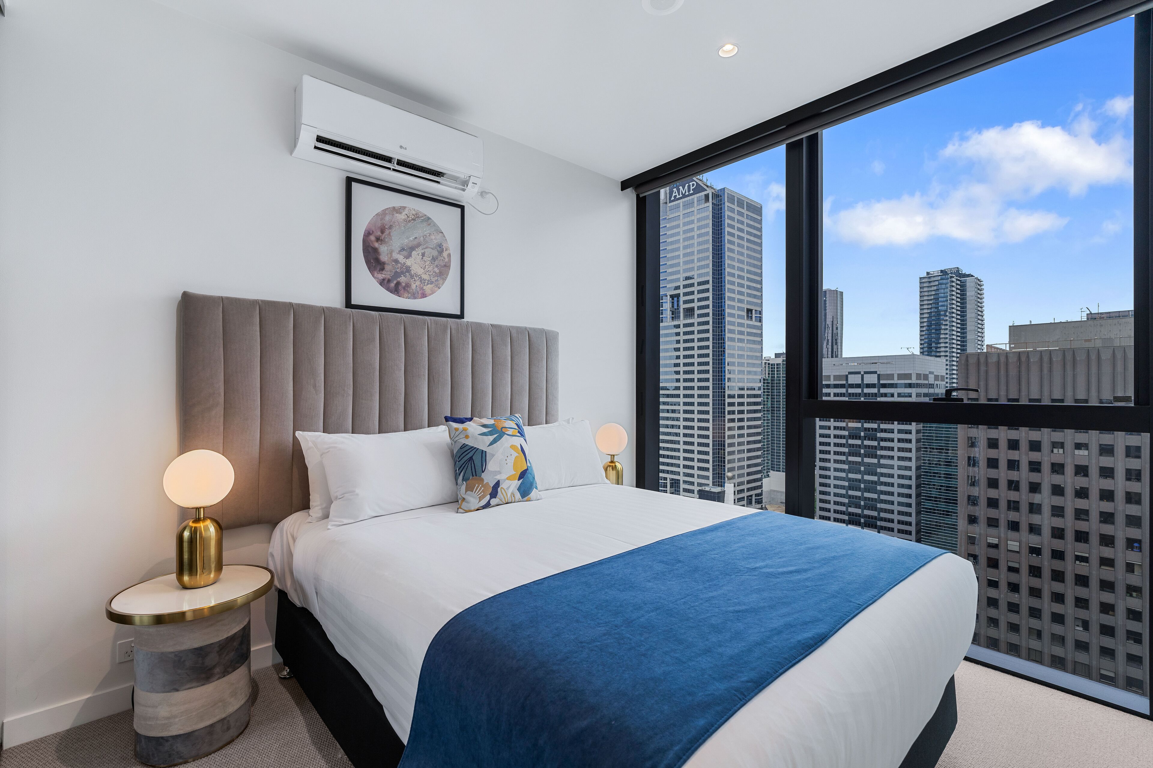 A modern hotel room with a large bed, a round bedside table, a wall-mounted air conditioner, and a city view through a large window.