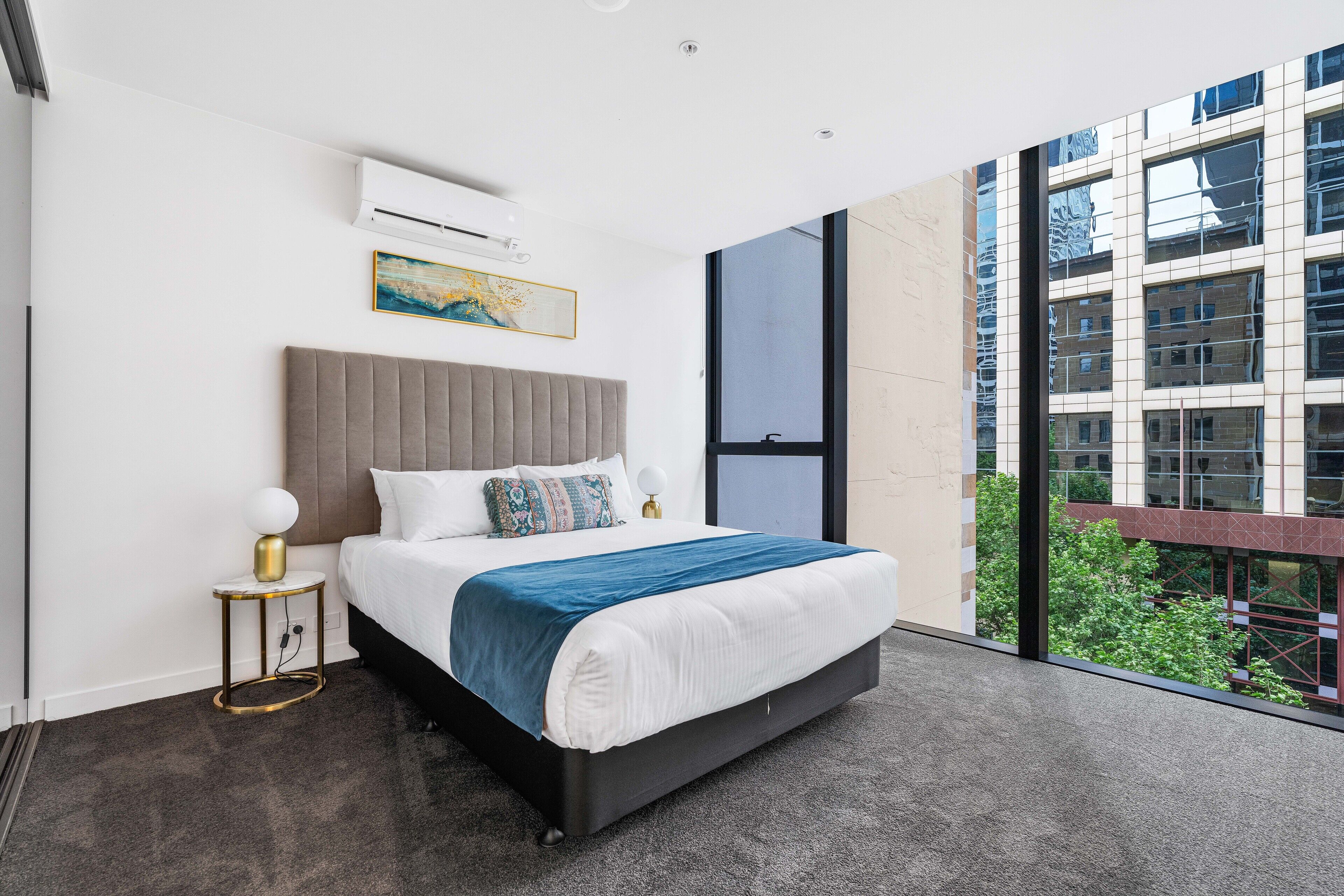 A modern hotel room with a large bed, a bedside table with a lamp, a wall-mounted air conditioner, a painting, and a view of a building and greenery outside.