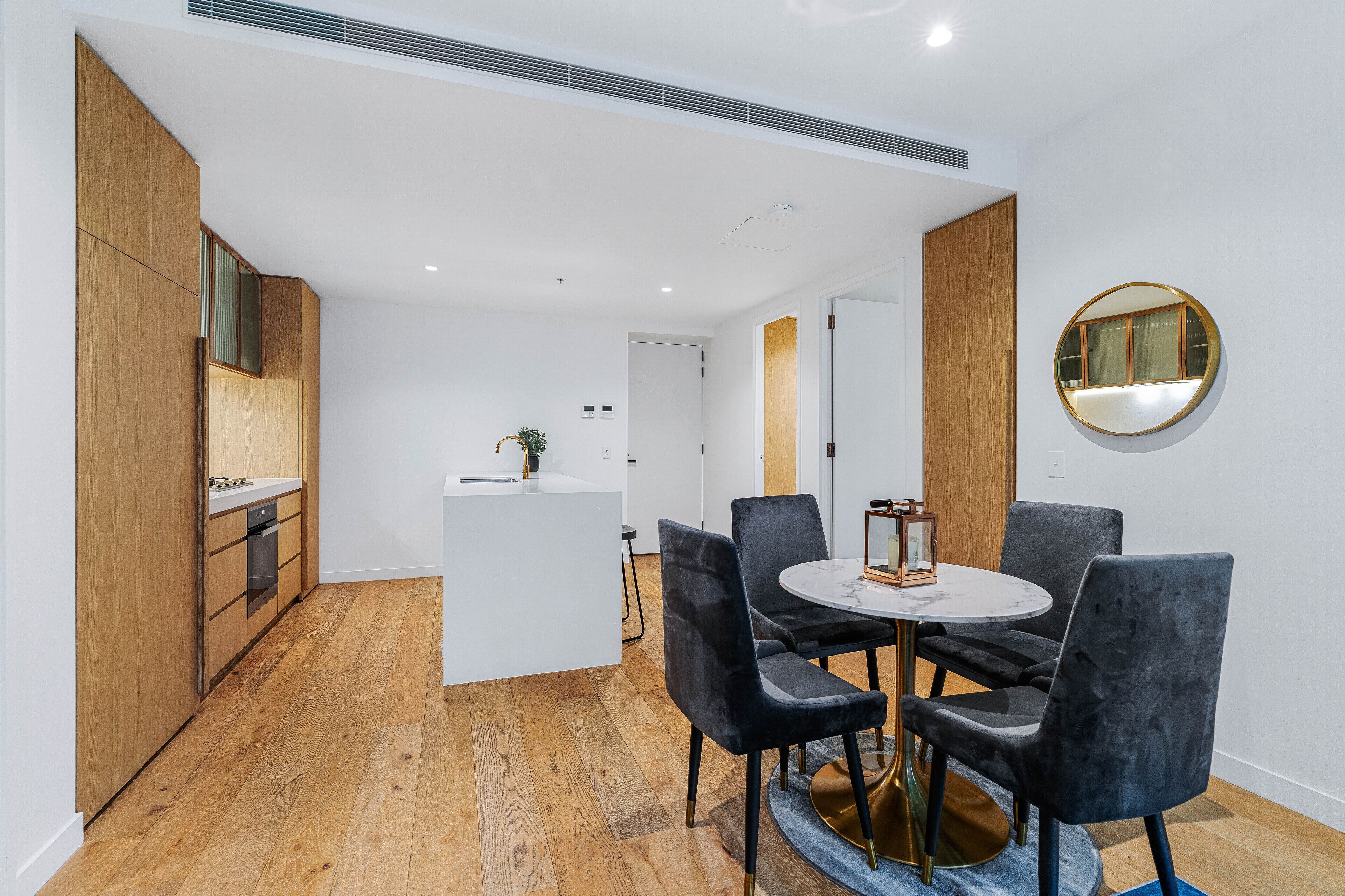 A modern kitchen with wooden cabinets and a white countertop, a dining area with a round table and four chairs, and a round mirror on the wall.