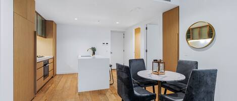 A modern kitchen with wooden cabinets and a white countertop, a dining area with a round table and four chairs, and a round mirror on the wall.
