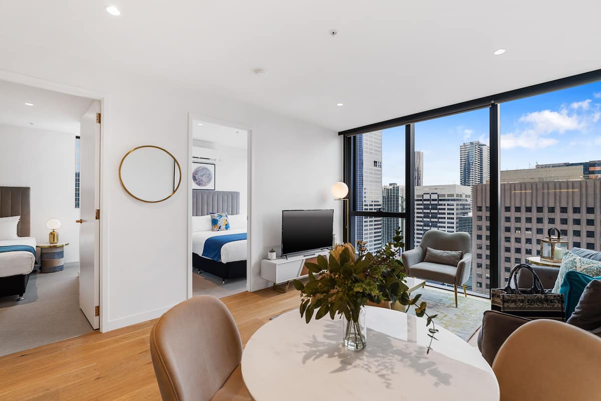 A modern hotel room with a large window, a flat-screen TV, a dining table, and a comfortable seating area.