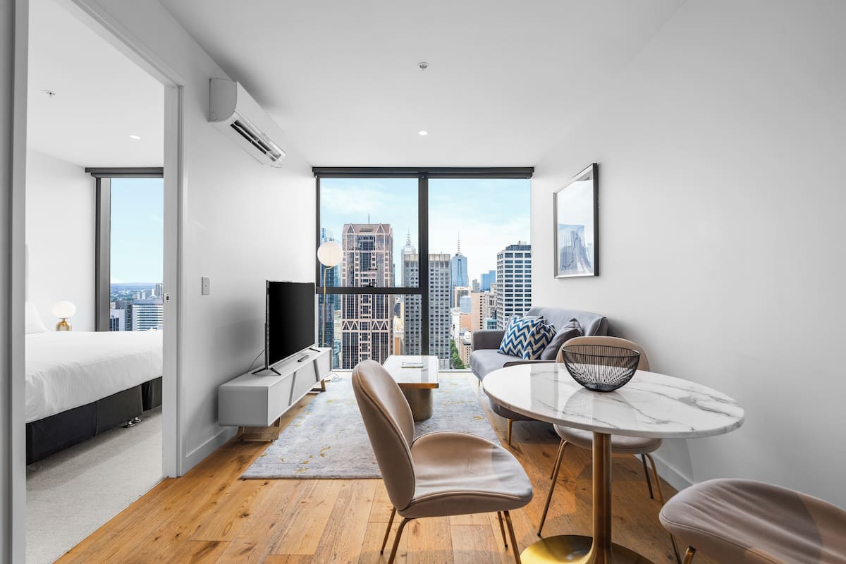 A modern hotel room with a large window, a bed, a sofa, a dining table, and a TV.