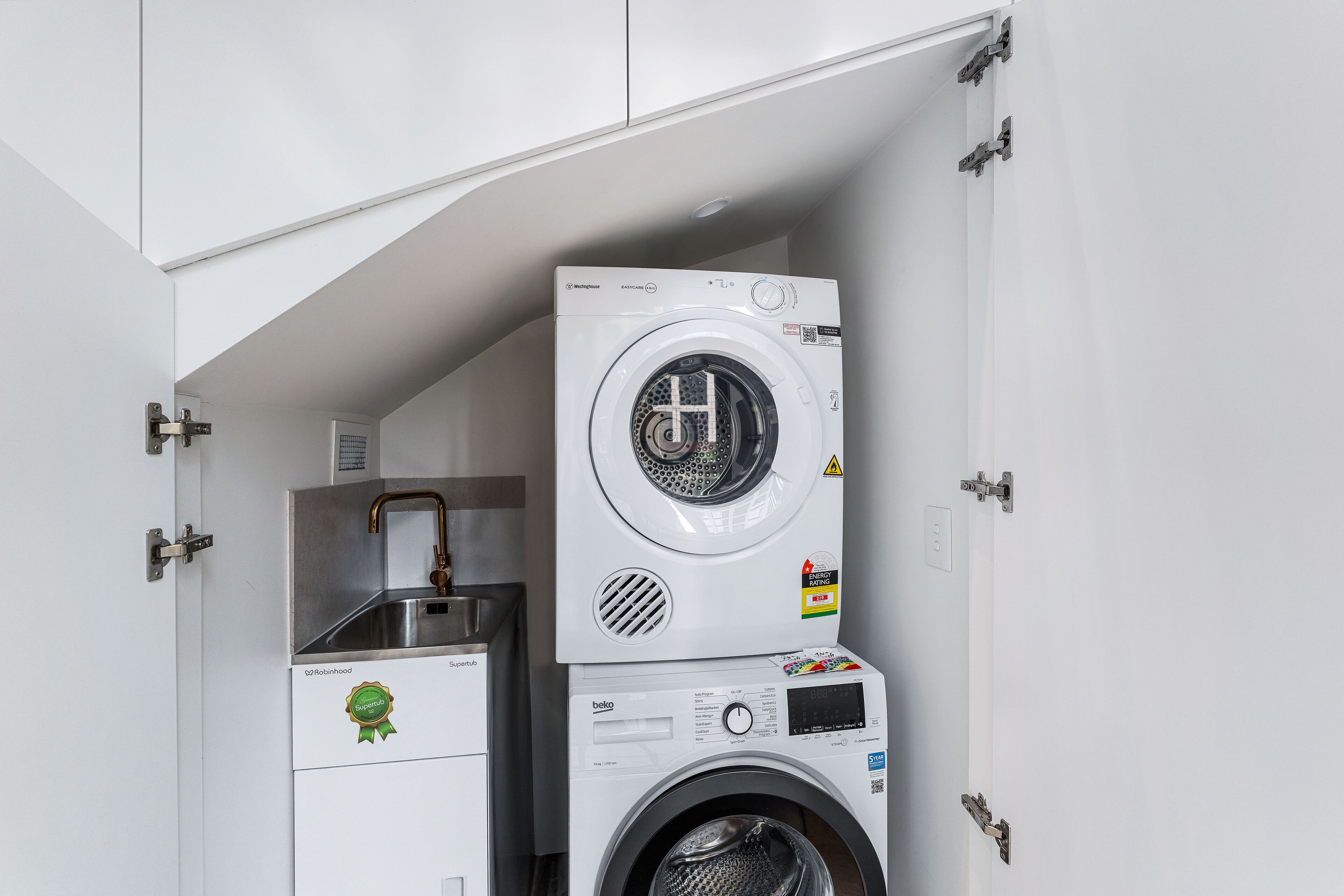 A compact laundry room with two stacked washing machines, a small sink, and a cabinet.