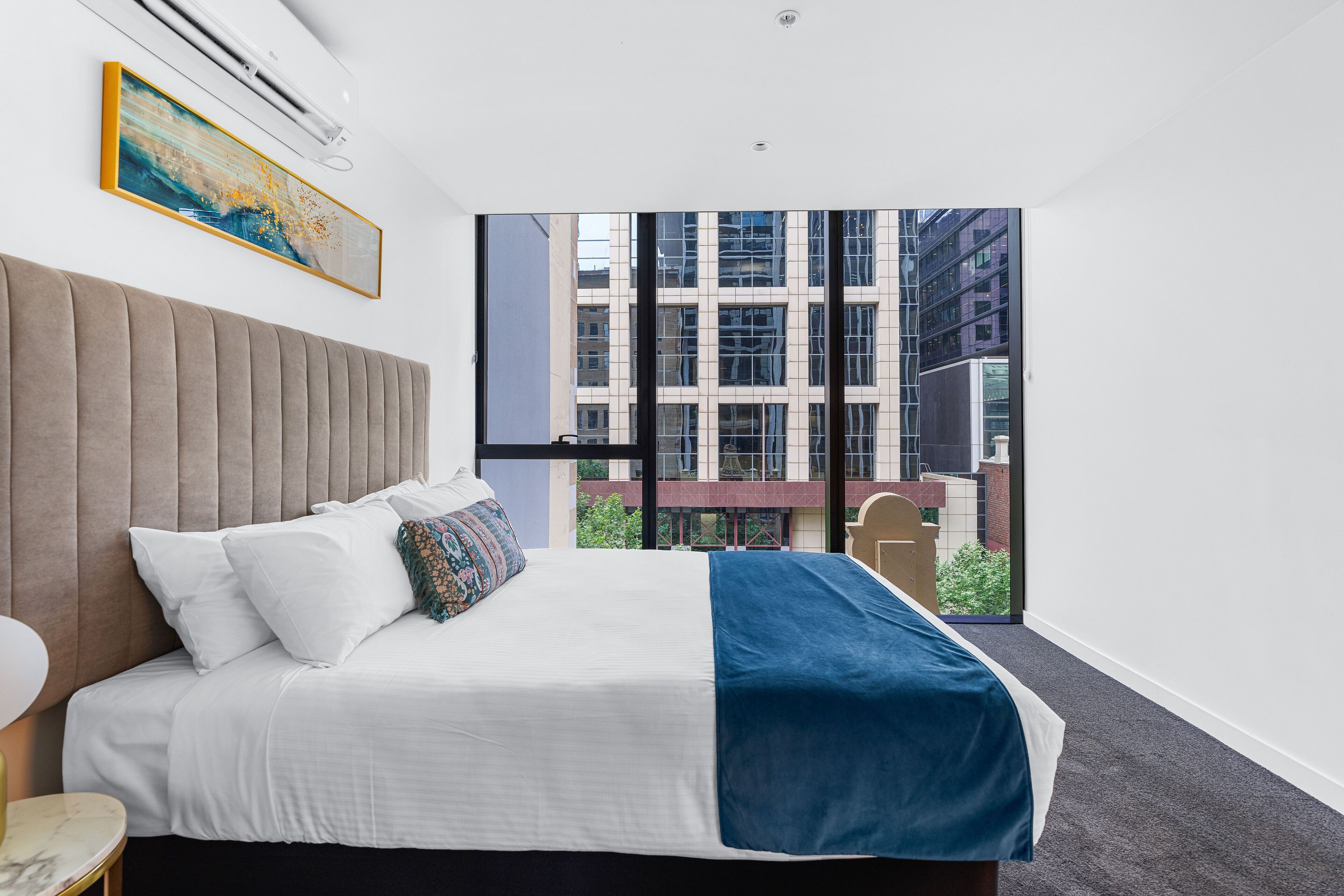 A modern hotel room with a large bed, a view of a cityscape through floor-to-ceiling windows, and a mounted air conditioner.