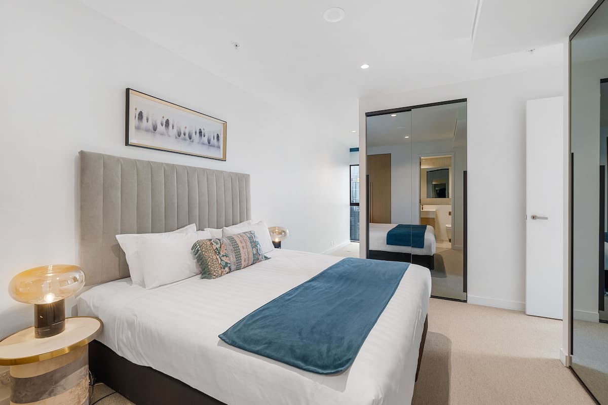 A modern hotel room with a large bed, a bedside table with a lamp, a framed picture on the wall, and a sliding door leading to another room.