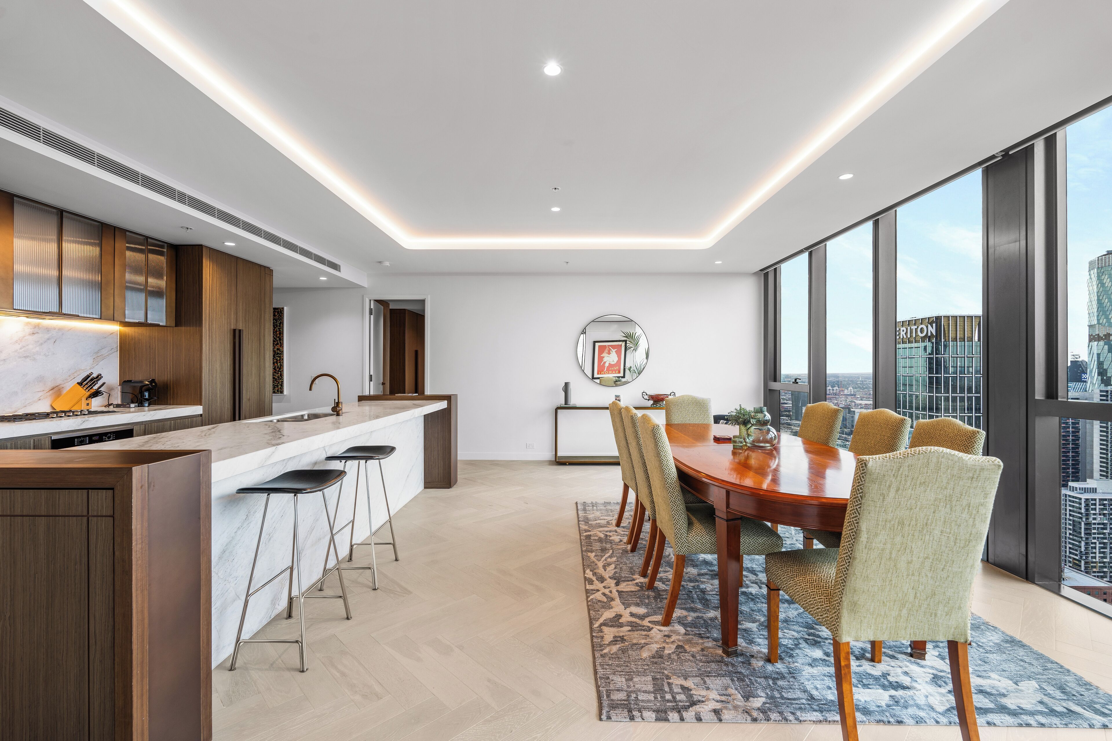 A modern kitchen with a marble countertop, wooden cabinets, and a bar area. There is a dining table with chairs and a view of the city through large windows.