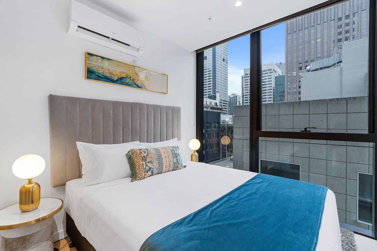 A modern hotel room with a large bed, a city view, and a mounted air conditioner.