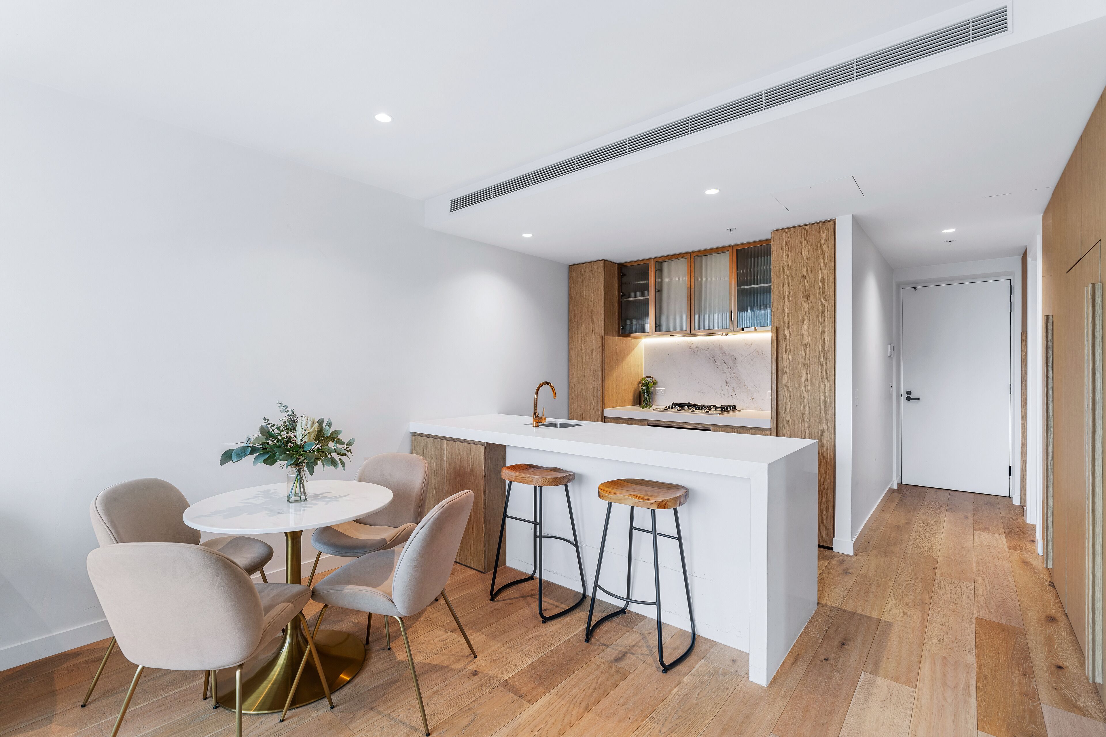 A modern kitchen with a white countertop, wooden cabinets, and a built-in oven. There are two bar stools and a round table with chairs.