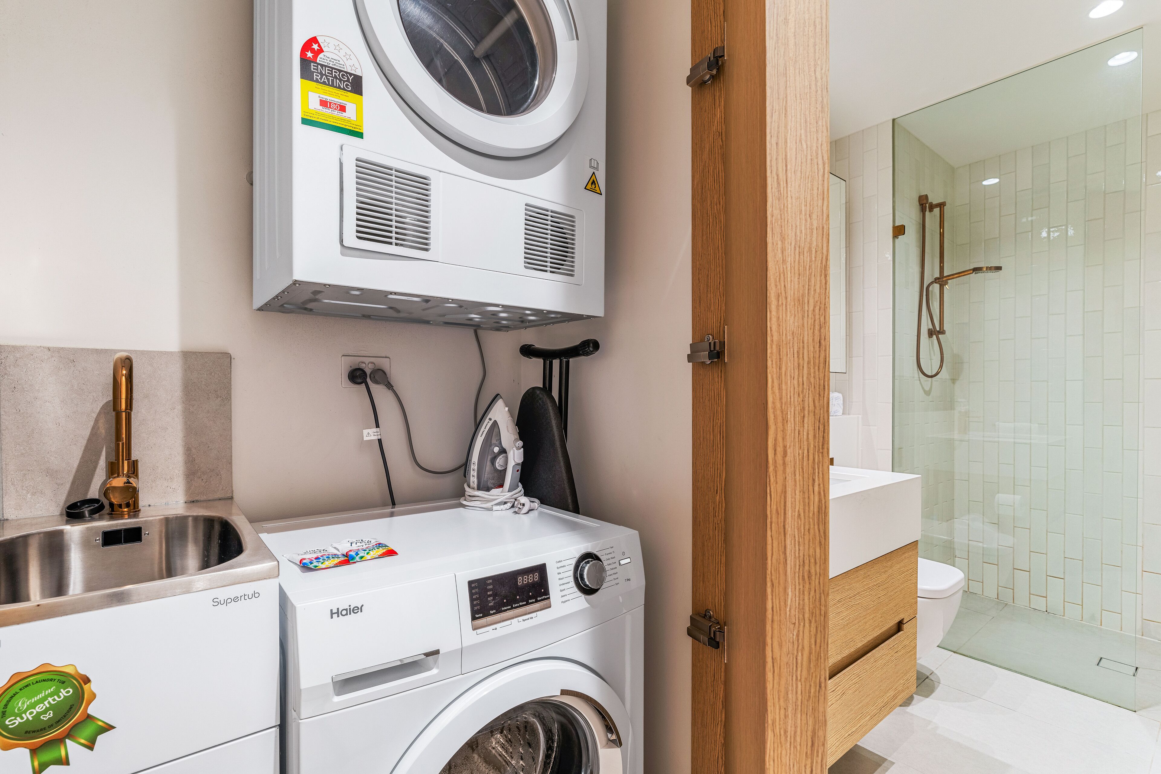 A compact laundry area with a front-loading washer and dryer, a stainless steel sink, and a glass-enclosed shower.
