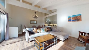 Ein Wohnzimmer mit einer Couch, einem Holztisch und einem Esstisch. Im Hintergrund befindet sich eine Küche, ein Chandelier und ein Gemälde an der Wand.