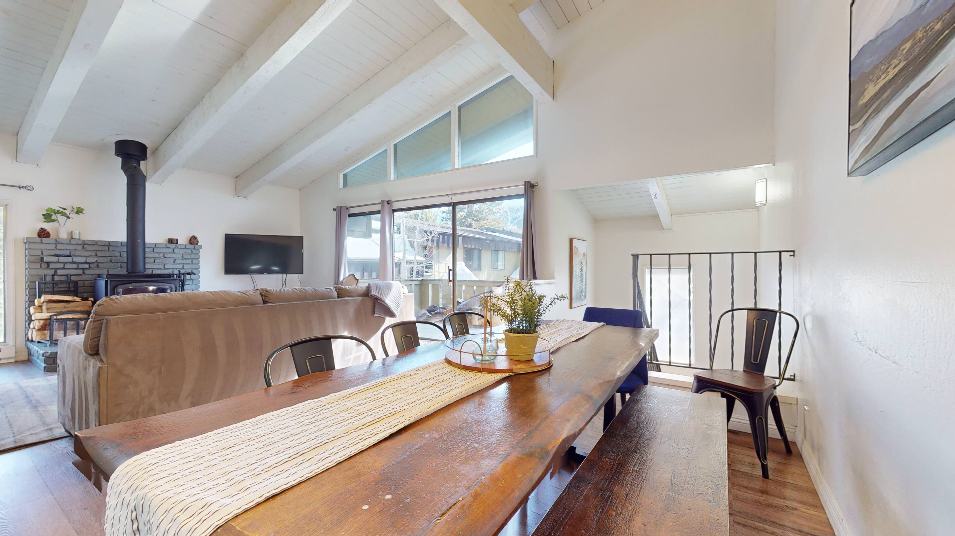 A spacious living room with a high ceiling, a stone fireplace, a large window, and a dining area with a wooden table and chairs.