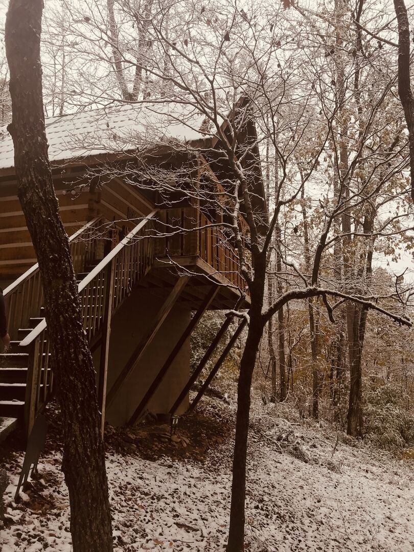 Une cabane en bois avec un escalier menant à une terrasse, entourée d’arbres et recouverte de neige.
