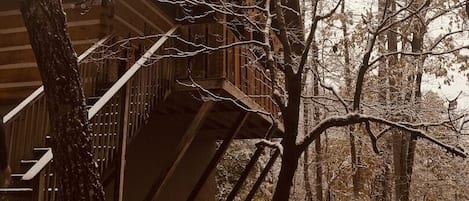 Una cabaña de madera con una escalera que lleva a una terraza, rodeada de árboles y cubierta de nieve.