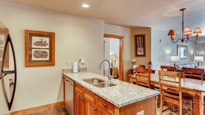 Una cocina con encimeras de granito, armarios de madera, un lavamanos de acero inoxidable y una zona de comedor con mesa y sillas de madera.