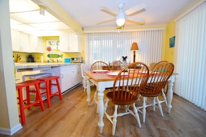 A dining area with a wooden table and chairs, a ceiling fan, and a kitchen area with white cabinets and red stools. - Thunder Island 112c 2 Bedroom Condo by RedAwning (Ocean City)
