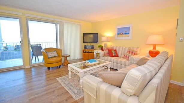 A living room with a sofa, armchair, coffee table, TV, and a balcony with a chair. - Thunder Island 112c 2 Bedroom Condo by RedAwning (Ocean City)
