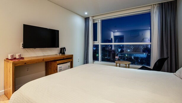 A hotel room with a large bed, a desk with a flat-screen TV, a coffee maker, and a view of a cityscape at night. - Dodubong Stay Hotel (Jeju City)