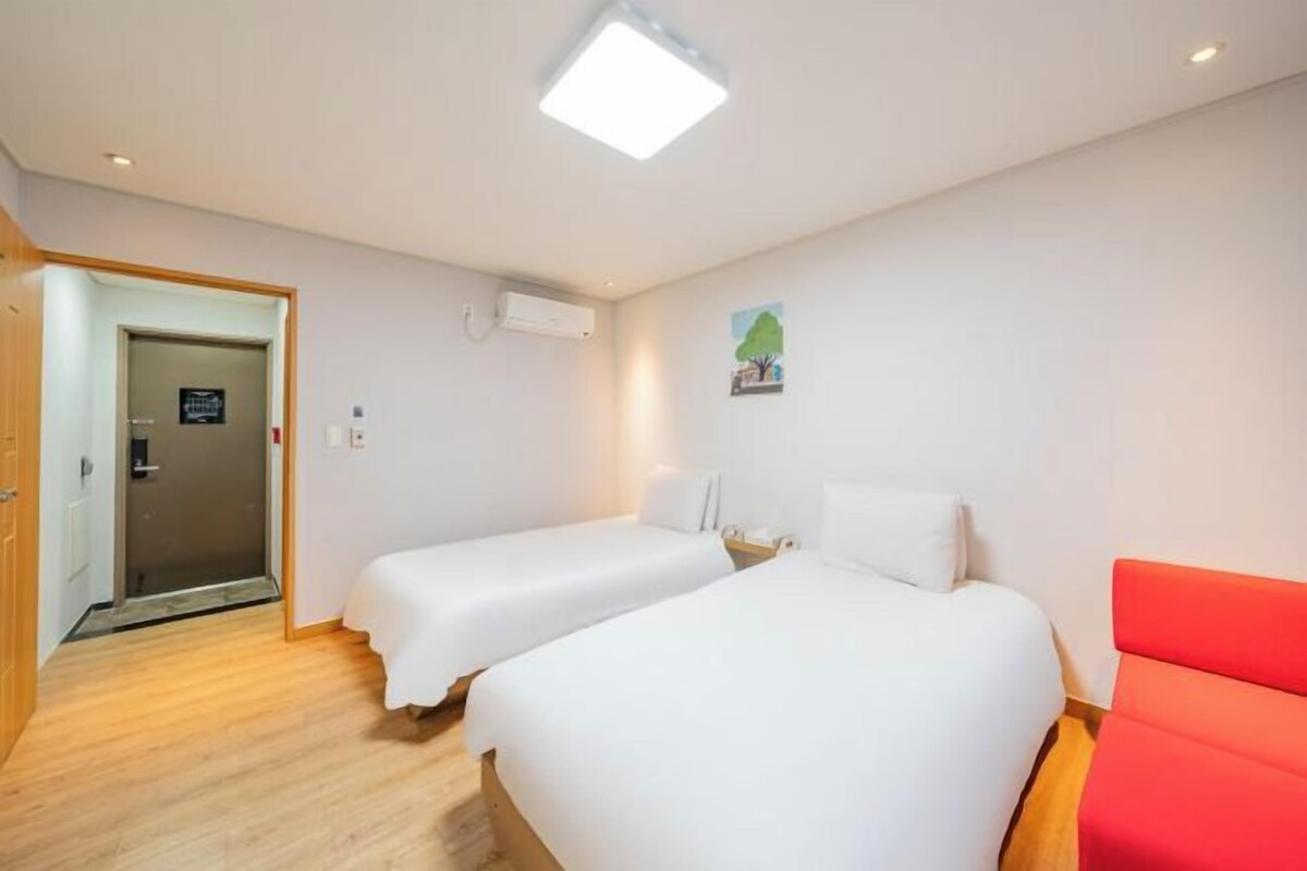 Ein Hotelzimmer mit zwei Betten, einem roten Sofa, einem Holzboden, einer weißen Decke und einer Tür zu einem weiteren Zimmer.