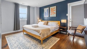 Een slaapkamer met een houten bed, een kamerkleed met patroon, een stoel en kunst aan de muur.