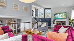Een ruime woonkamer met een open haard, een grote televisie en een trap naar een hoger gelegen verdieping.