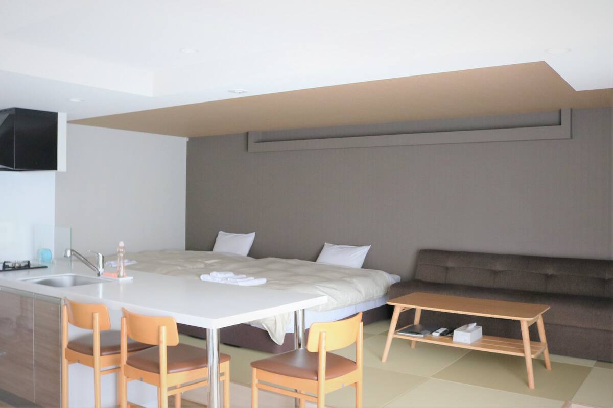 A modern, compact room with a kitchenette, a bed with a headboard, a sofa, and two wooden chairs.