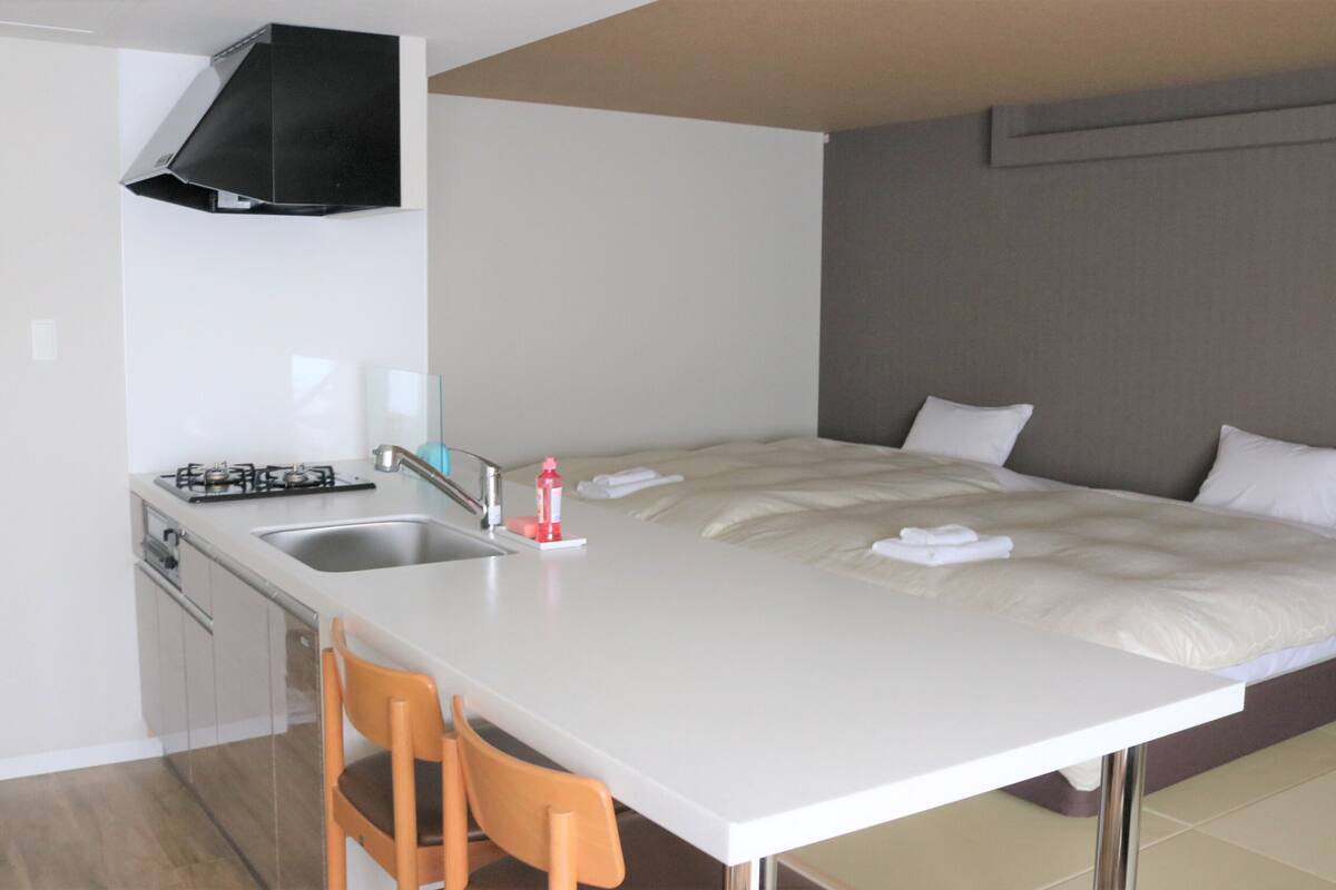 A compact hotel room with a kitchenette, a bed with white linens, and a small dining area with a chair.