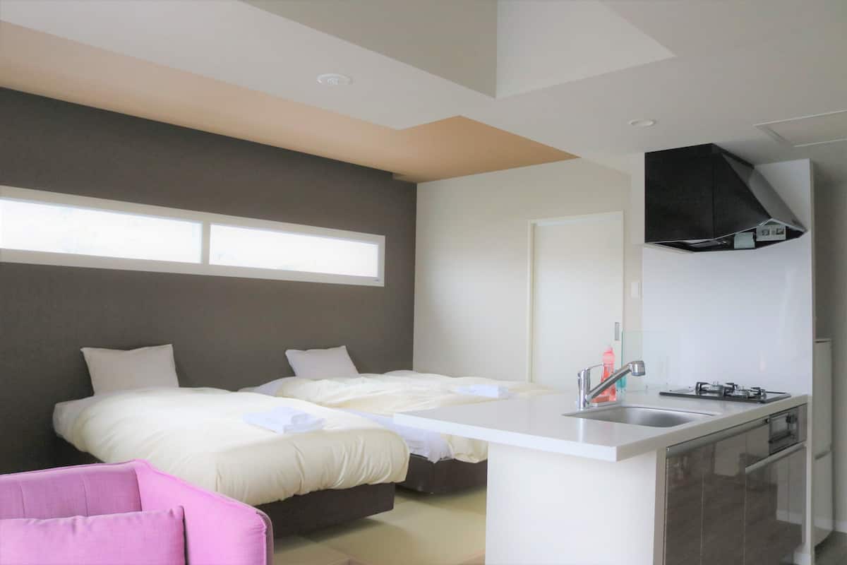 A modern hotel room with a bed, a small kitchenette, and a window providing natural light.