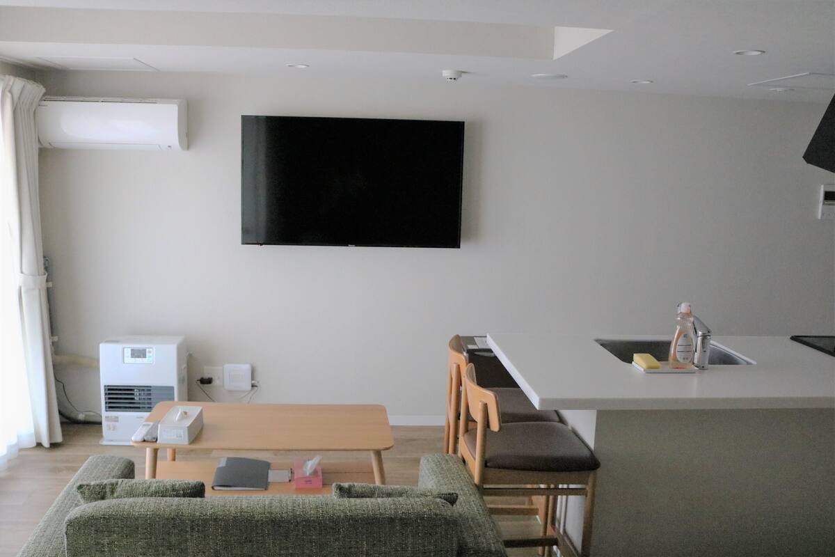 A modern living room with a flat-screen TV, a wooden dining table, and a kitchen area with a sink and bar.