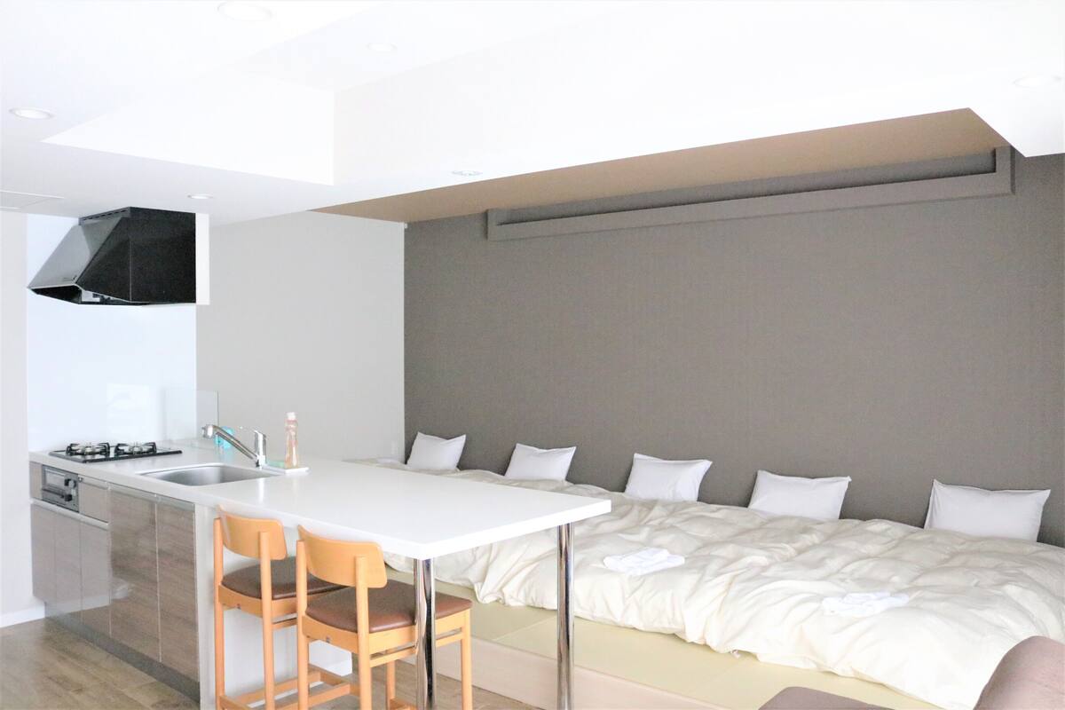 A modern hotel room with a kitchenette, a bed with a large headboard, and a seating area with two chairs.