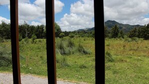 A view through a window with a natural landscape outside, featuring grassy fields, trees, and distant mountains under a partly cloudy sky. - Stunning Cabin w/360º view of Guatemala Highlands (Tecpán Guatemala)