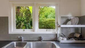 A kitchen with a double sink, a window with a view of greenery, and a dish rack with plates and cups. - Taman Indrakila (Ubud)