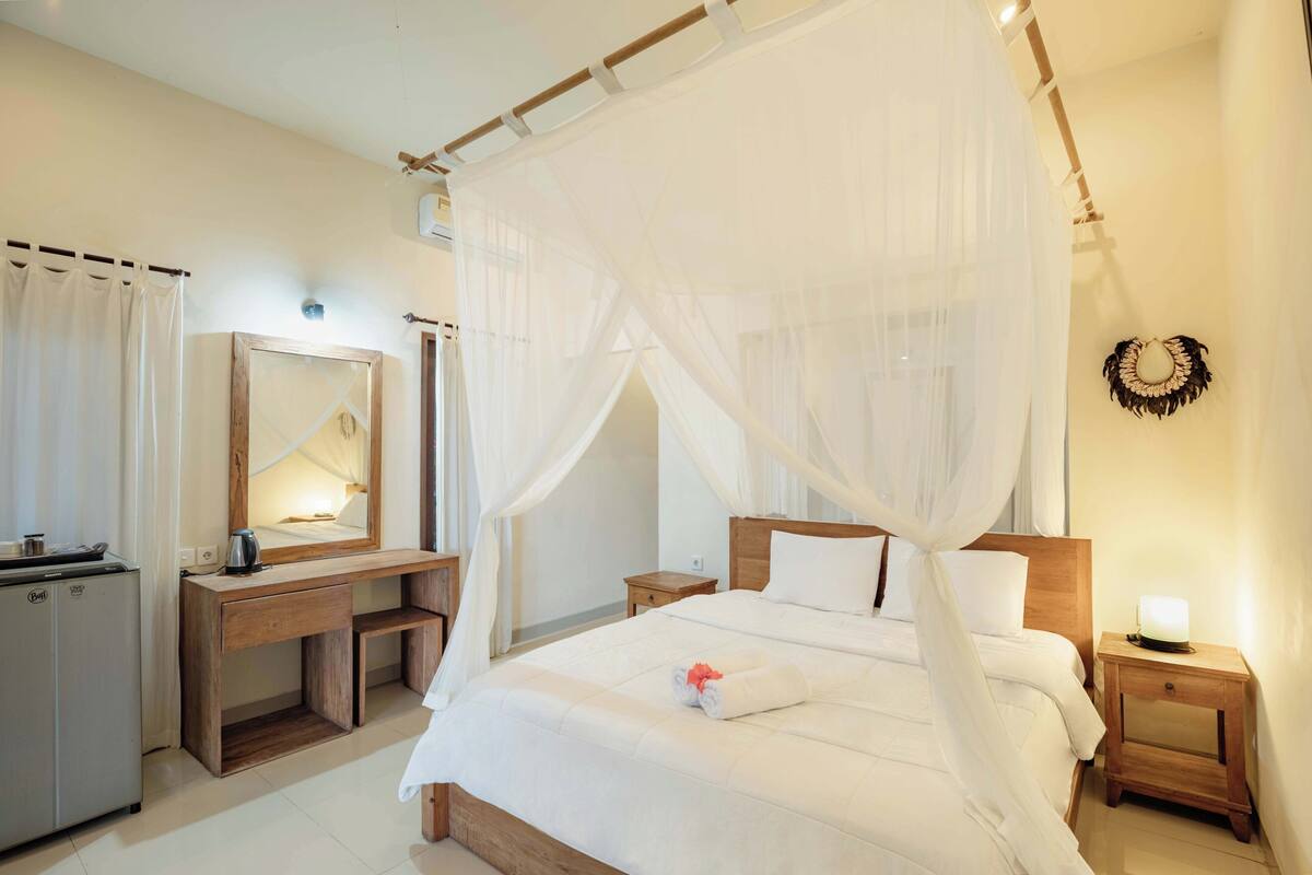 A bedroom with a four-poster bed, a wooden desk, a mirror, and a nightstand with a lamp.