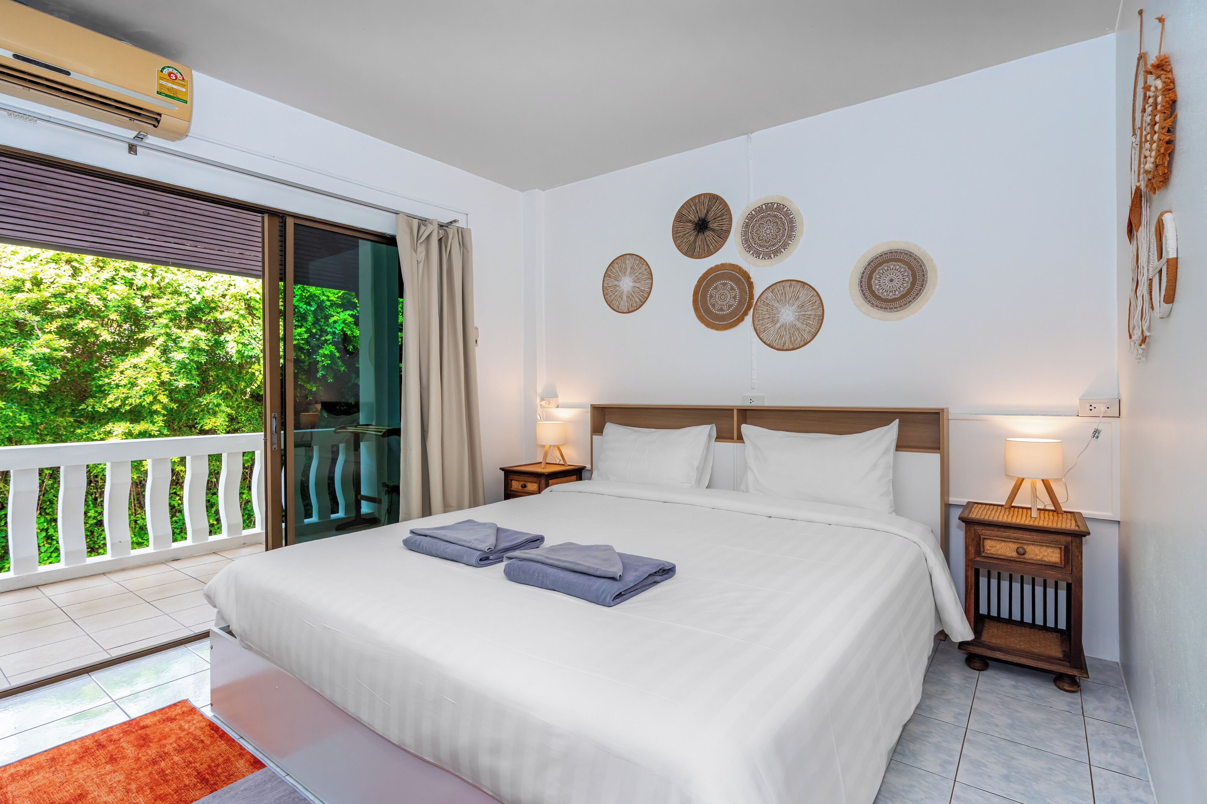 A hotel room with a large bed, two bedside lamps, a wall with decorative plates, a balcony with a view of greenery, and an air conditioning unit.