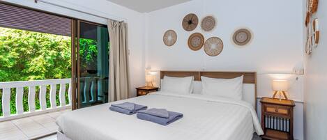 A hotel room with a large bed, two bedside lamps, a wall with decorative plates, a balcony with a view of greenery, and an air conditioning unit.