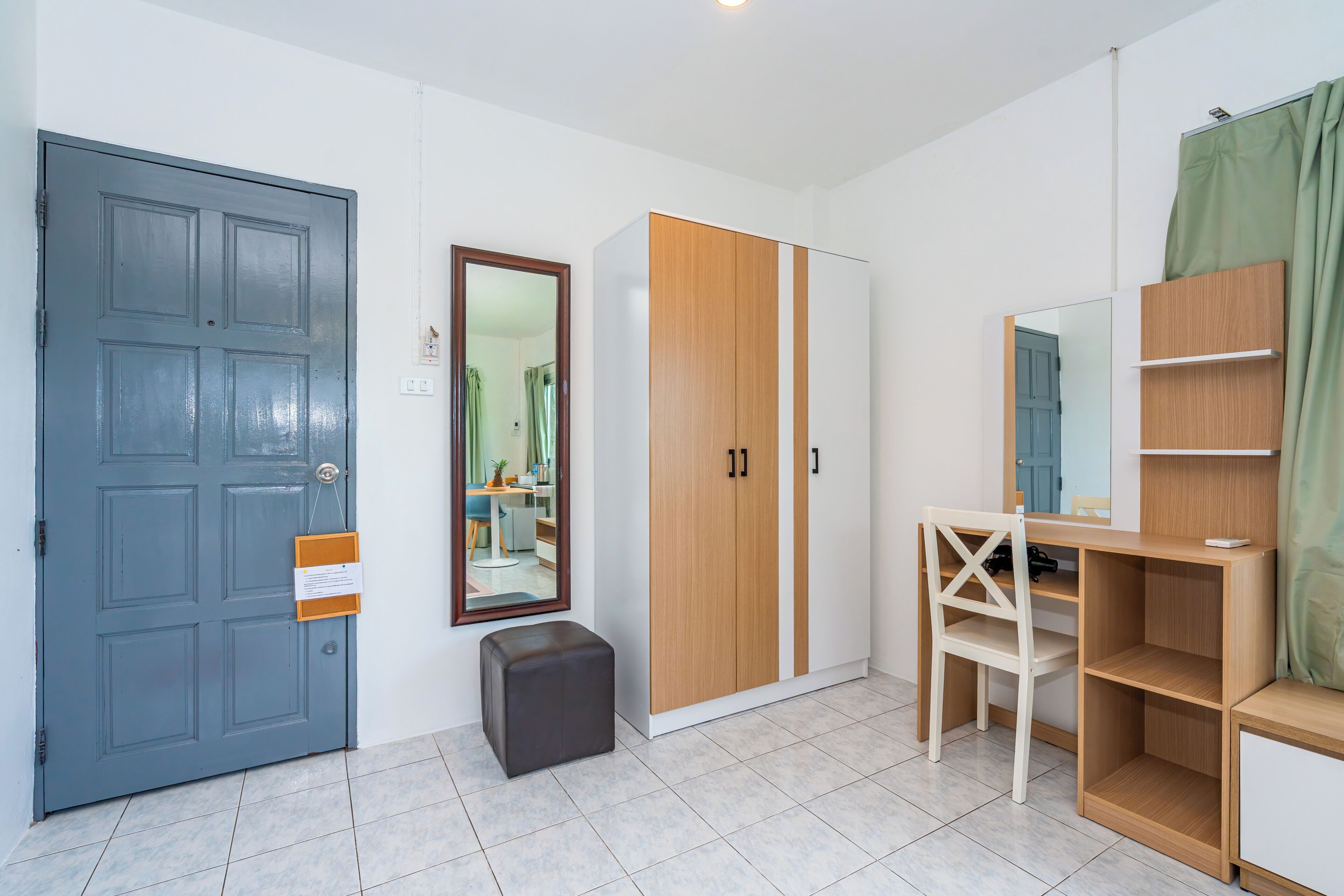 A room with a grey door, a wooden wardrobe, a mirror, a small stool, and a desk with shelves.