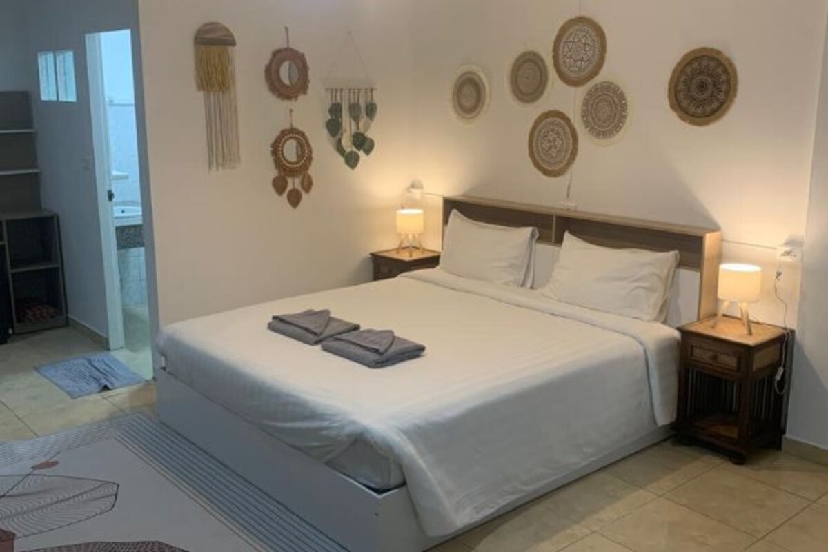 Una cama bien hecha con cubrecama y almohadas blancas, dos mesitas de noche con lámparas y adornos en la pared.